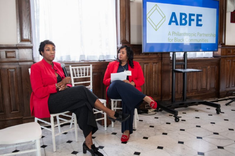Two Black women in red blazers and black pants sitting next to a screen displaying the ABFE logo