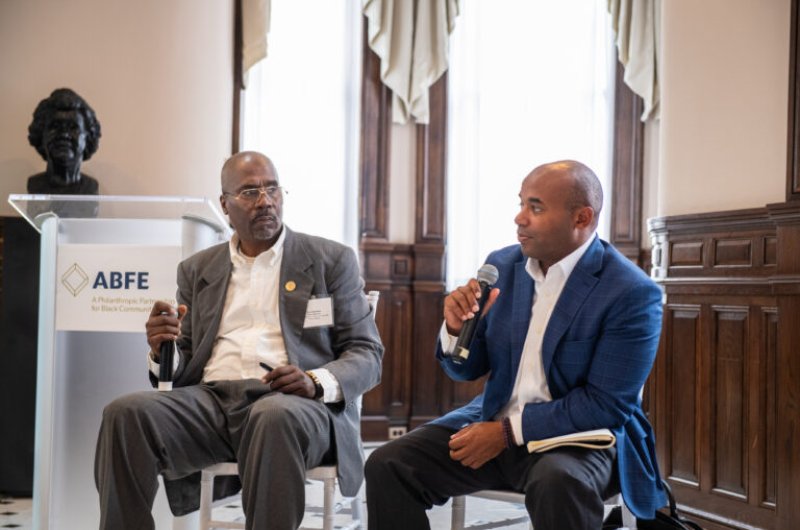 Two Black men in suits seated next to each other holding microphones in a discussion